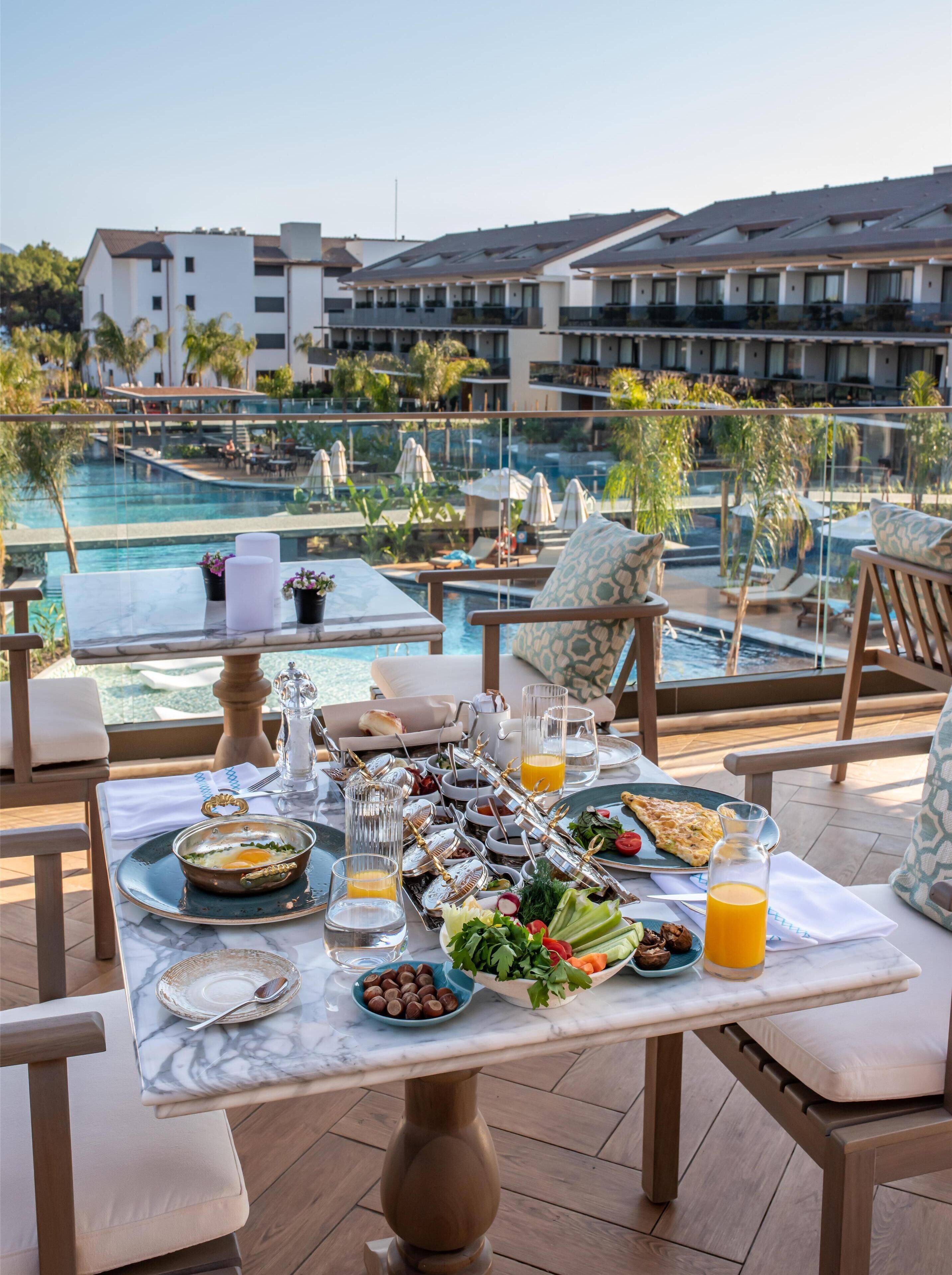 Ein Esstisch gedeckt mit Frühstücksangeboten, Blick auf einen Pool und eine luxuriöse Hotelanlage.