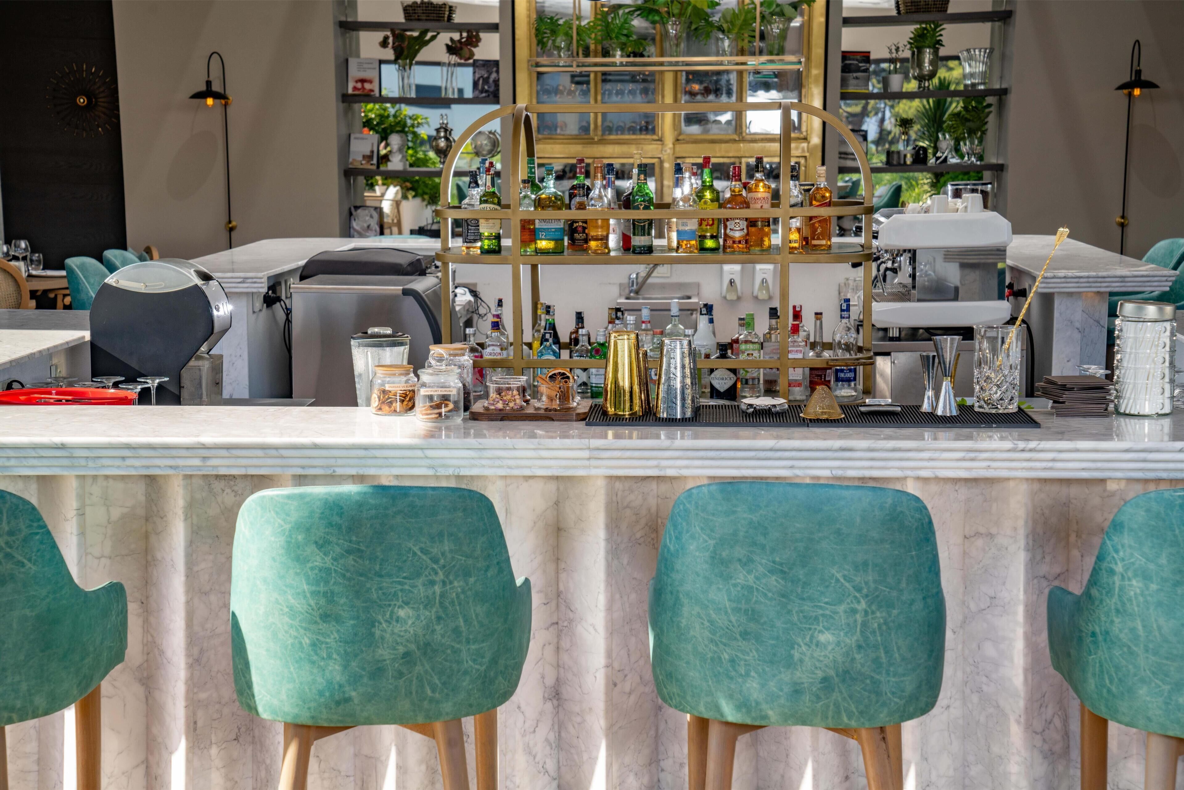 A bar area with a marble countertop, green upholstered chairs, and a gold-framed liquor display.