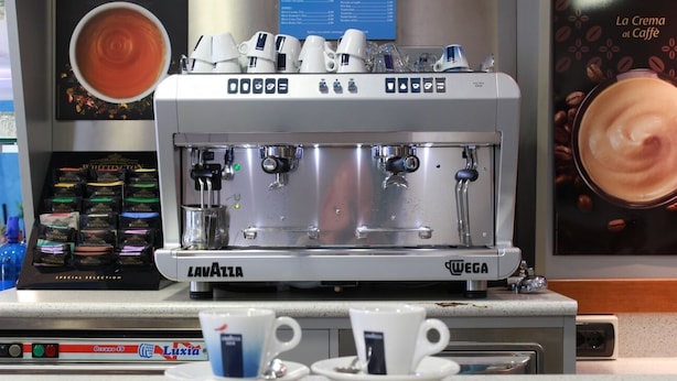 A coffee machine with multiple portafilters and a digital display, cups on a saucer, and a menu board in the background. - Hotel Nettuno (Montecatini Terme)