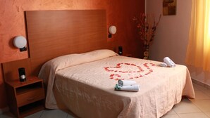 A neatly made bed with a heart-shaped decoration on the bedspread, a nightstand with a coffee maker, and a vase with branches on the wall. - Hotel Nettuno (Montecatini Terme)