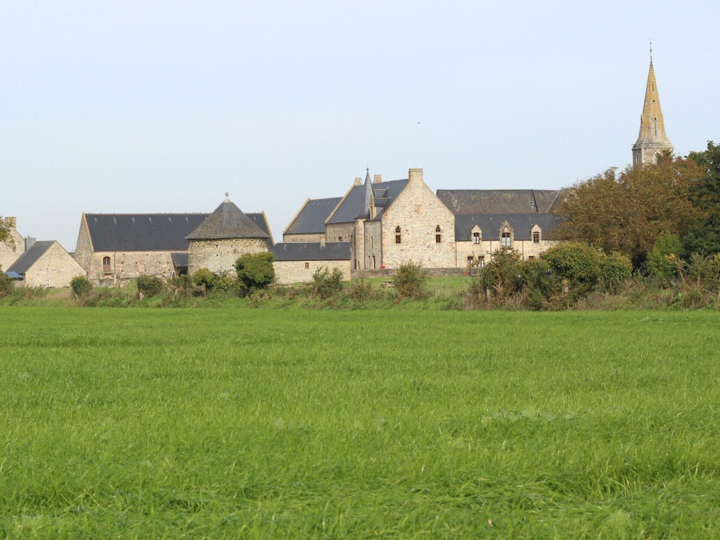 Un bâtiment historique en pierre, composé de plusieurs structures, dont une église surmontée d’une flèche, situé dans un paysage rural avec un grand champ verdoyant au premier plan.