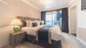 A bedroom with a large bed, a bedside table with a lamp, a view of the outside through a window, and a door to the right. - The Fisherbeck (Ambleside)