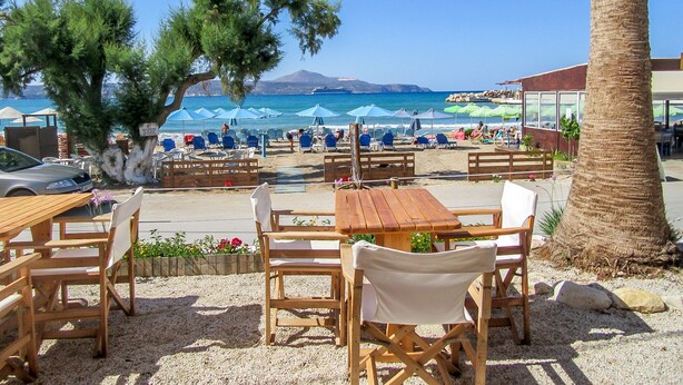 Een café aan het strand met houten tafels en stoelen, parasols en uitzicht op zee en bergen.