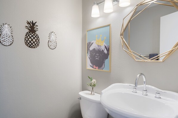 A bathroom with a toilet, sink, and wall-mounted mirror. There are three pineapple decorations on the wall and a framed picture of a dog with a crown.