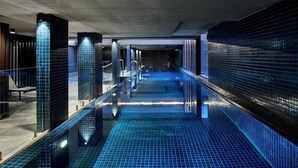 Una piscina interior con suelo y paredes de azulejos, rodeada por barandillas de vidrio y pilares iluminados.