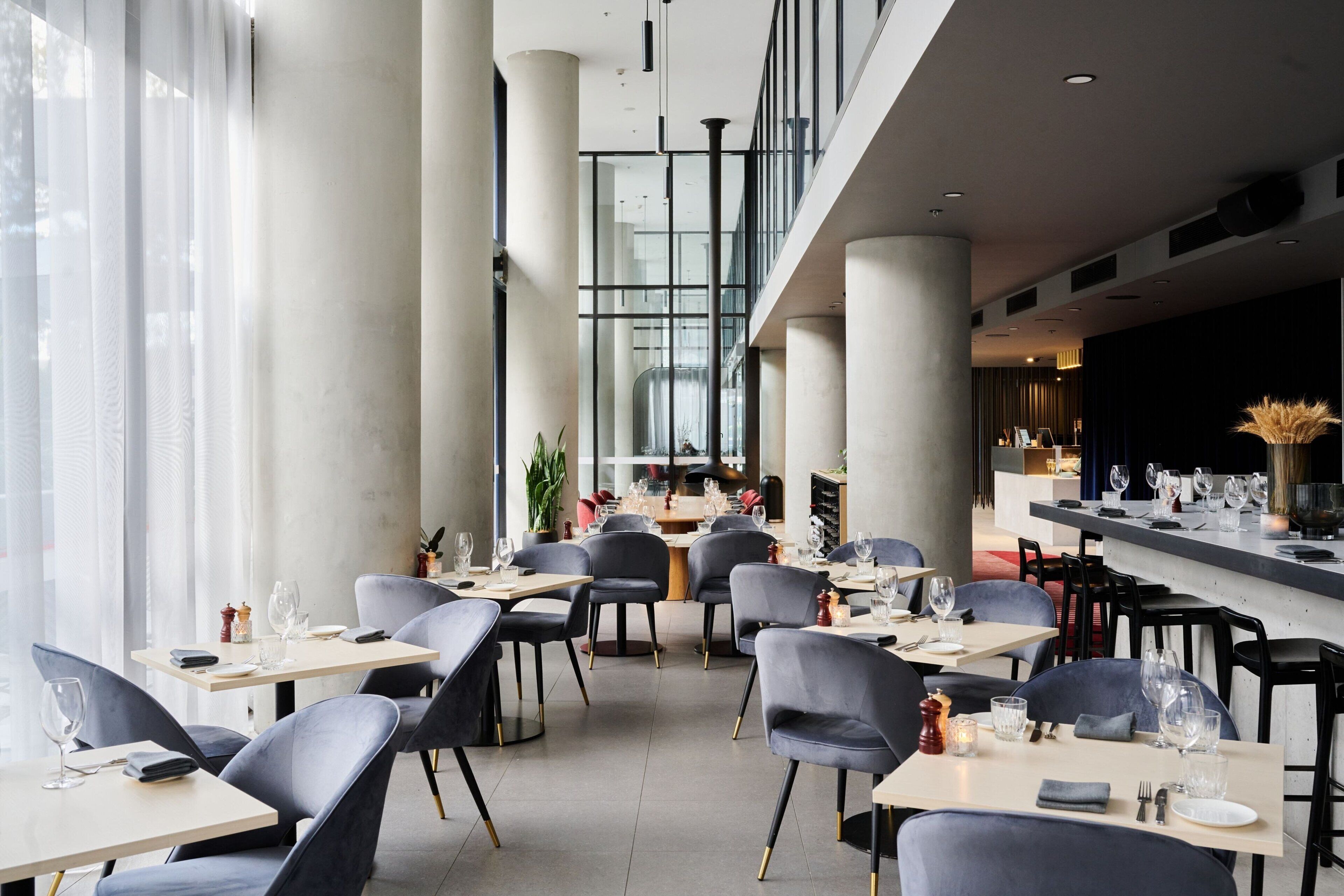 A modern restaurant with high ceilings, large windows, and a mezzanine level. Tables are set with wine glasses and cutlery.