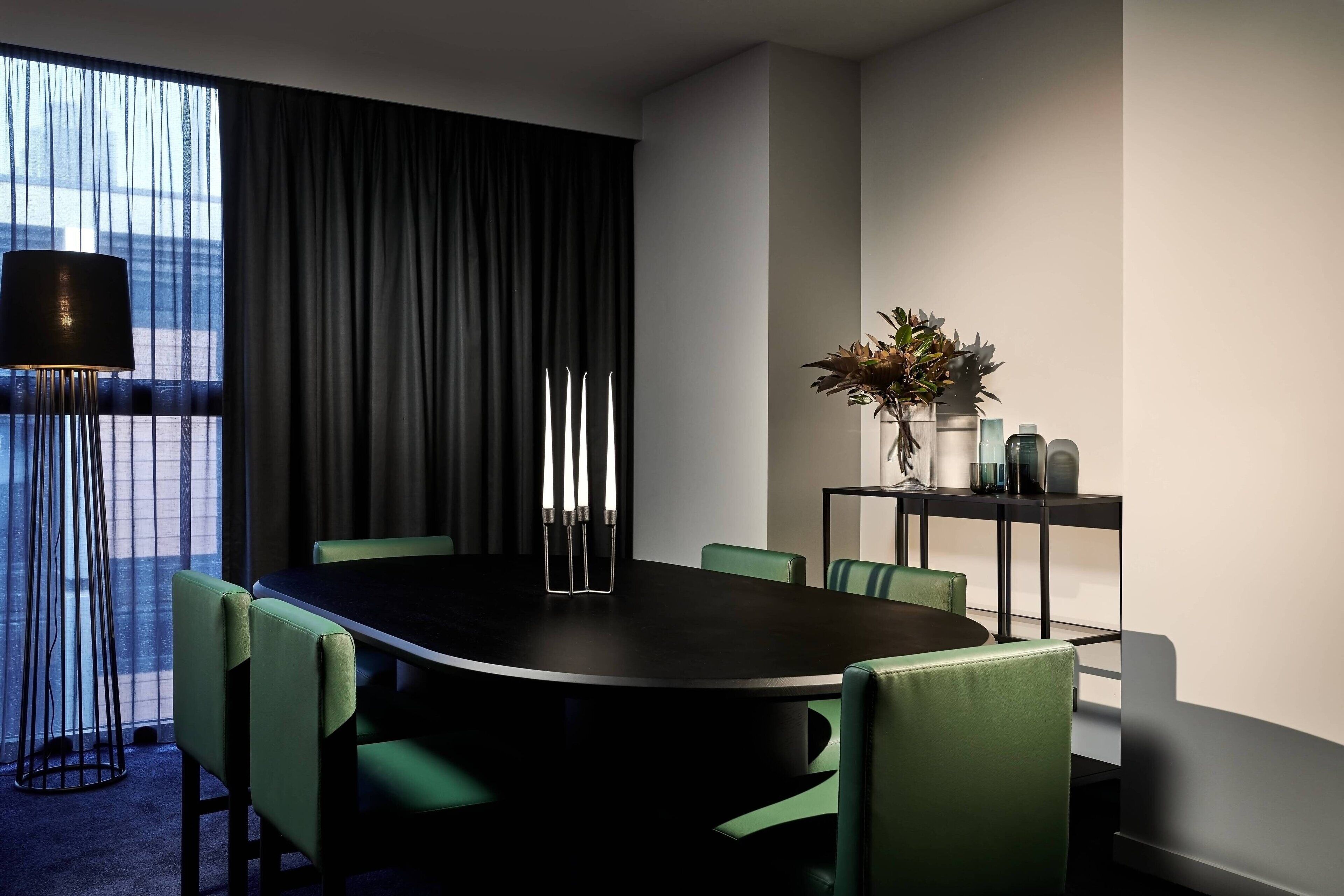 Un salón de conferencias con una mesa oscura, sillas verdes, una lámpara de pie alta y una planta sobre una mesita.