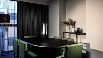 Un salón de conferencias con una mesa oscura, sillas verdes, una lámpara de pie alta y una planta sobre una mesita.