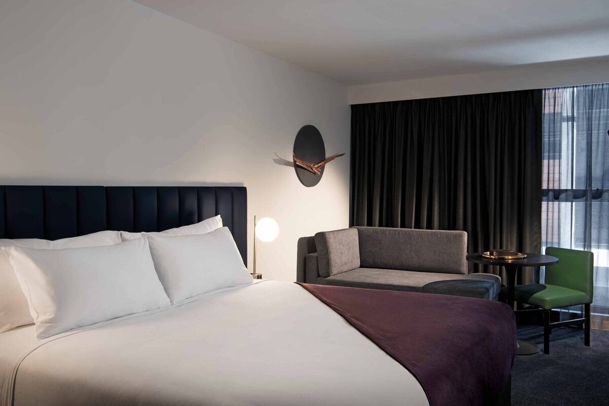 A hotel room with a large bed, a dark headboard, a wall-mounted clock, a side table with a lamp, a green chair, and a view of the city through large windows.