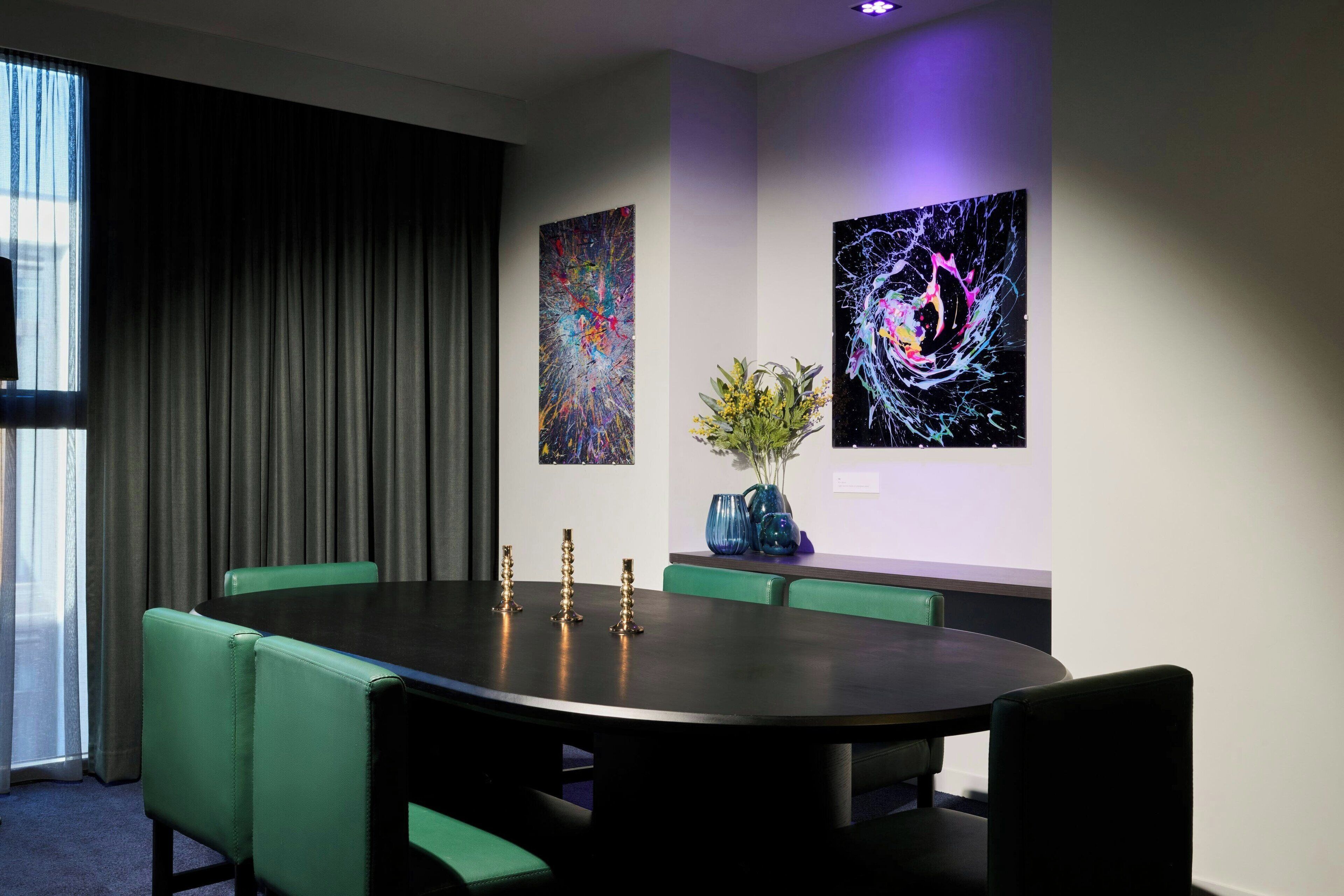 Une salle de conférence avec une table ronde, des chaises recouvertes d’un tissu vert et deux tableaux abstraits accrochés aux murs.