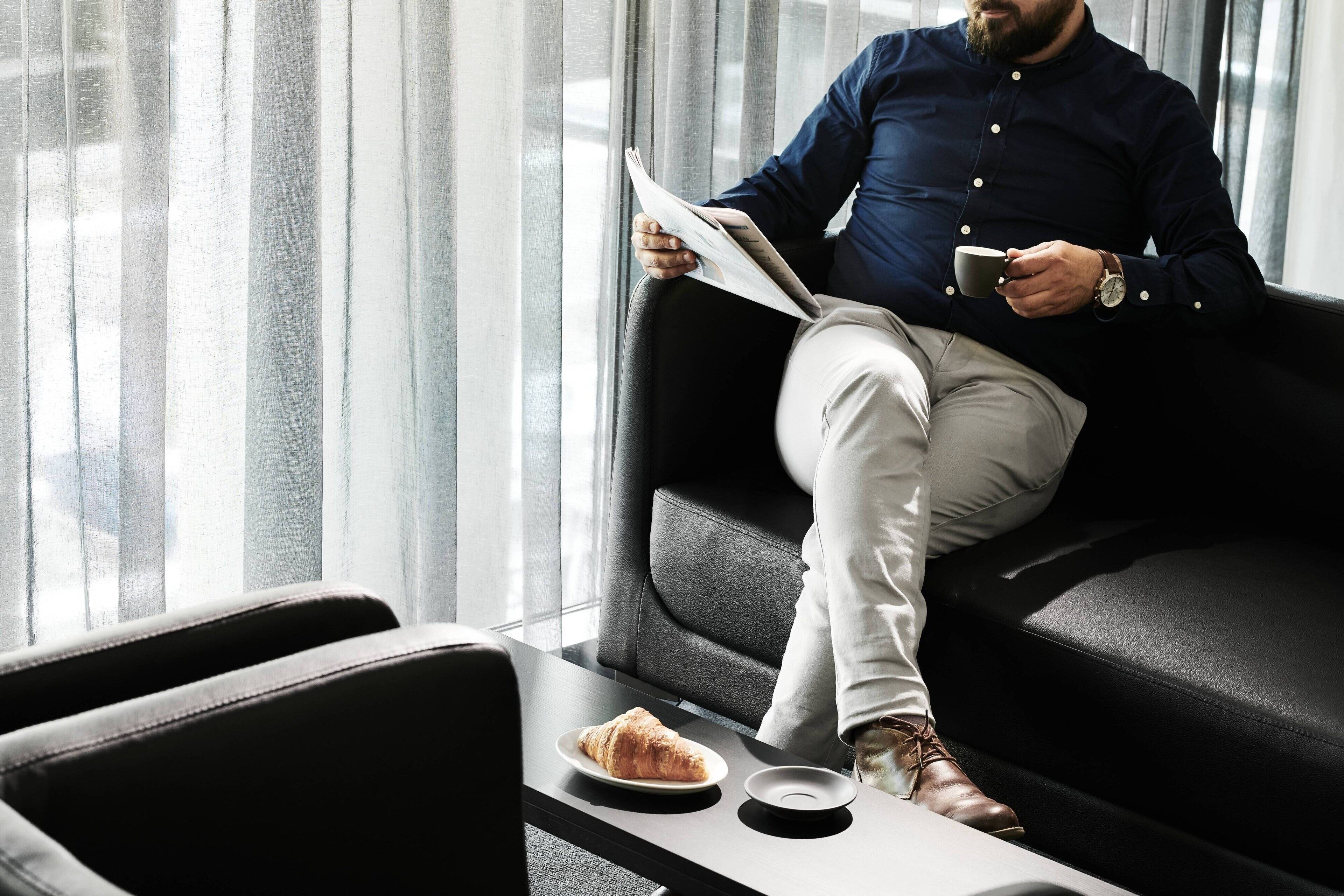 A man sitting on a black leather couch reading a newspaper, with a croissant and a cup on a table beside him.