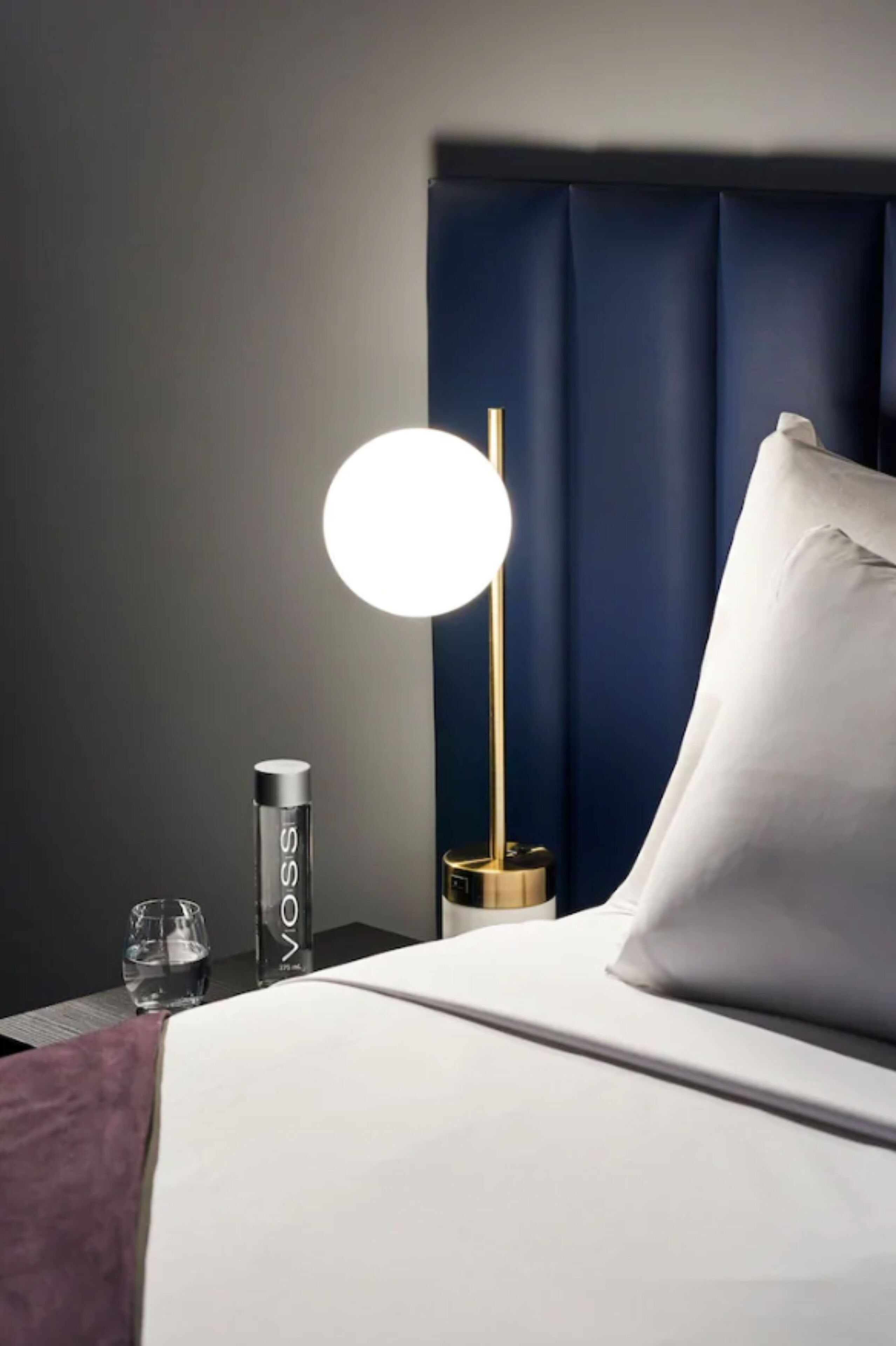 A neatly made bed with a white pillow and a round table lamp with a gold base.