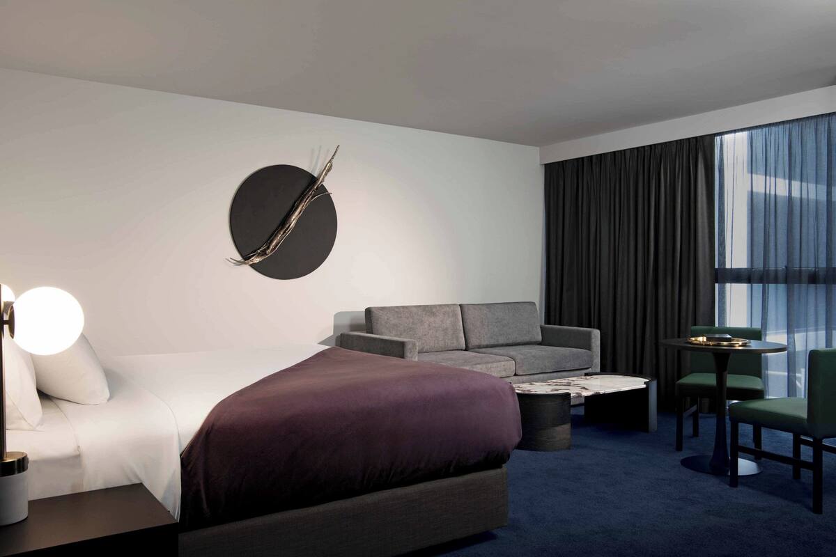 A modern hotel room with a large bed, a sofa, a coffee table, and a dining table with chairs.