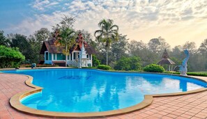 A swimming pool with a tiled edge, surrounded by a landscaped garden, a house with a distinctive roof, and a statue.