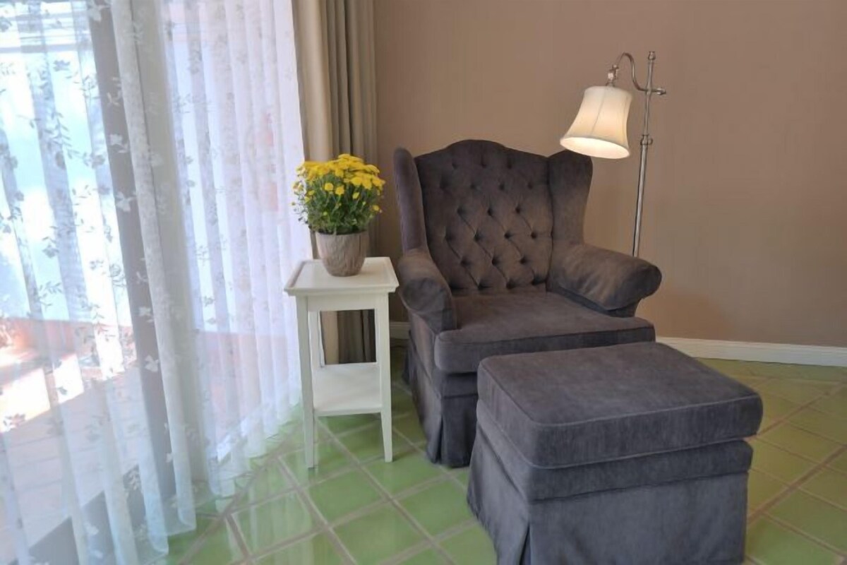 A room with a grey upholstered armchair and ottoman, a small white side table with a potted plant, and a floor lamp.