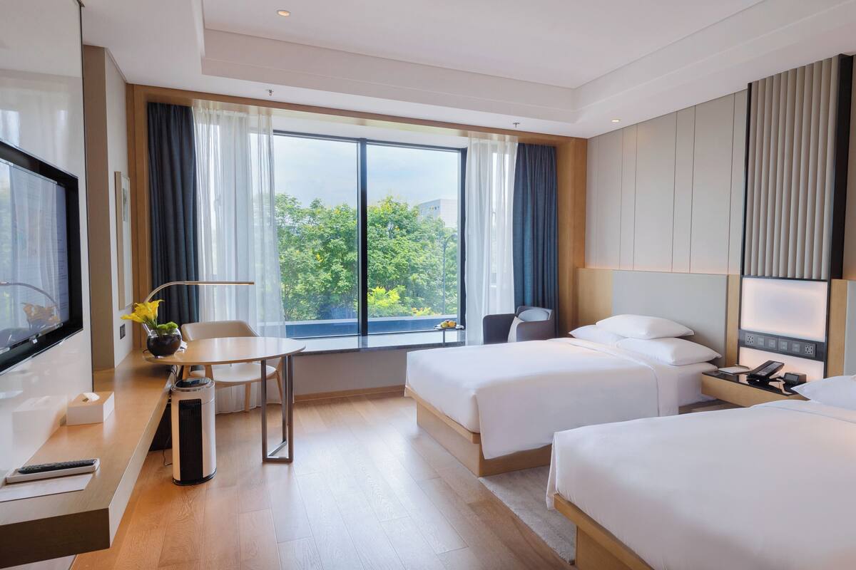 Habitación de hotel con dos camas, un escritorio con silla, televisión, ventana con cortinas y vista a árboles.