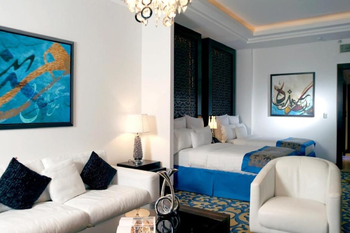 A hotel room with a bed, a sofa, a chair, a lamp, and artwork on the walls.
