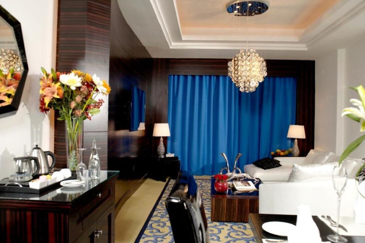 A hotel room with a dining area, a sofa, a chandelier, and a large window with curtains.