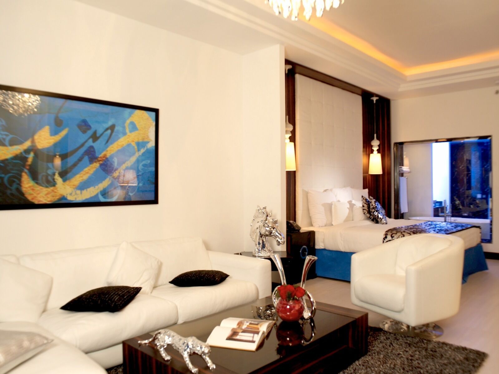 Habitación de hotel moderna con un sofá blanco, una mesa de centro con objetos decorativos, una cama con ropa de cama blanca y un cuadro abstracto grande en la pared.