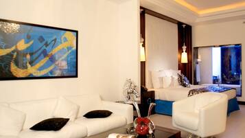 Habitación de hotel moderna con un sofá blanco, una mesa de centro con objetos decorativos, una cama con ropa de cama blanca y un cuadro abstracto grande en la pared.