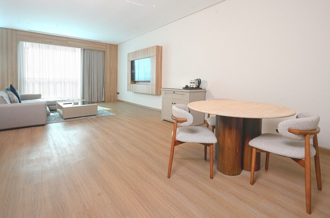 Uma sala de estar moderna com um sofá, uma mesa de centro e uma mesa de jantar com cadeiras. Há uma televisão de tela plana na parede e uma pequena área de cozinha com uma cafeteira.