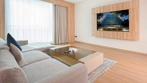 Una sala de estar moderna con un televisor de pantalla plana grande montado en la pared, un sofá color beige con cojines, una mesa de centro de vidrio y un ventanal con cortinas translúcidas.
