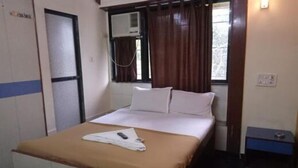 A single bed with a brown bedspread, a white pillow, and a remote control on the bed. There is a window with brown curtains, an air conditioning unit, and a small table with a lamp next to the bed. - Summerland Guest House (Mumbai)