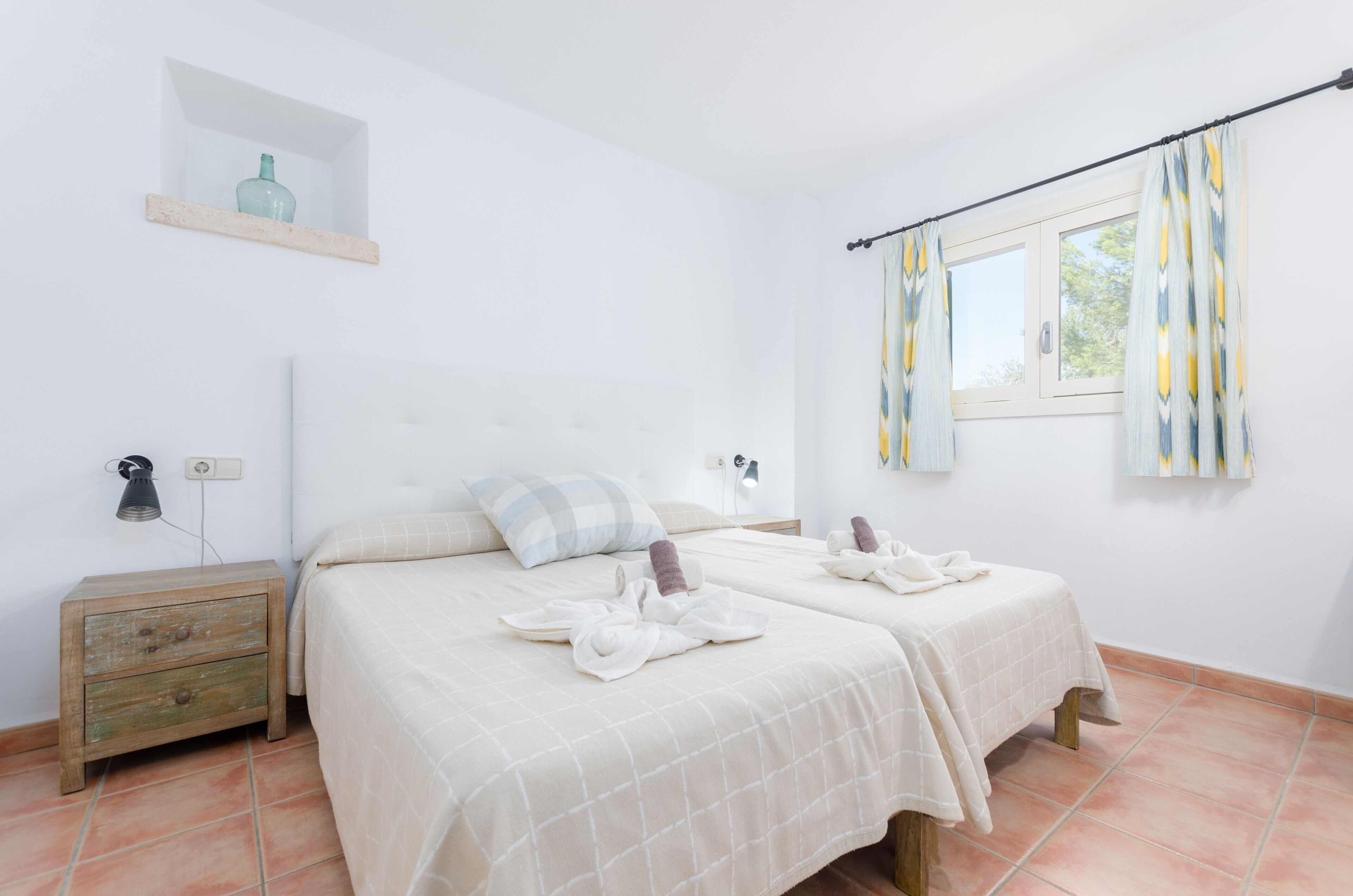 Une chambre avec deux lits, une table de chevet, une étagère avec une bouteille et une fenêtre avec des rideaux.