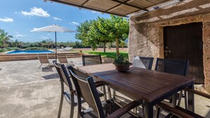 Un patio exterior con mesa y sillas de madera, una planta en maceta y vistas a una piscina y árboles.
