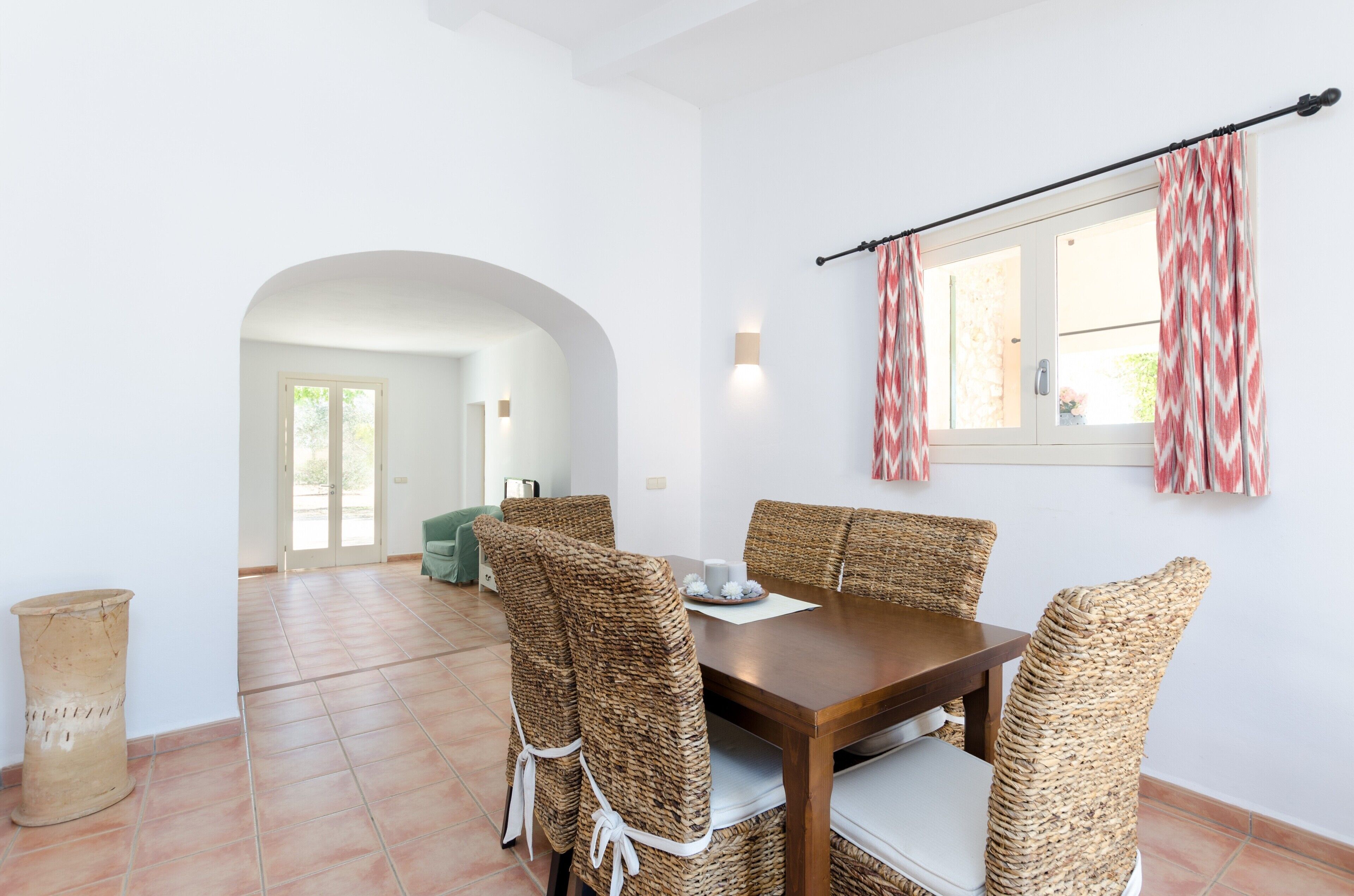 A dining area with a wooden table and woven chairs, a window with patterned curtains, and a tiled floor.