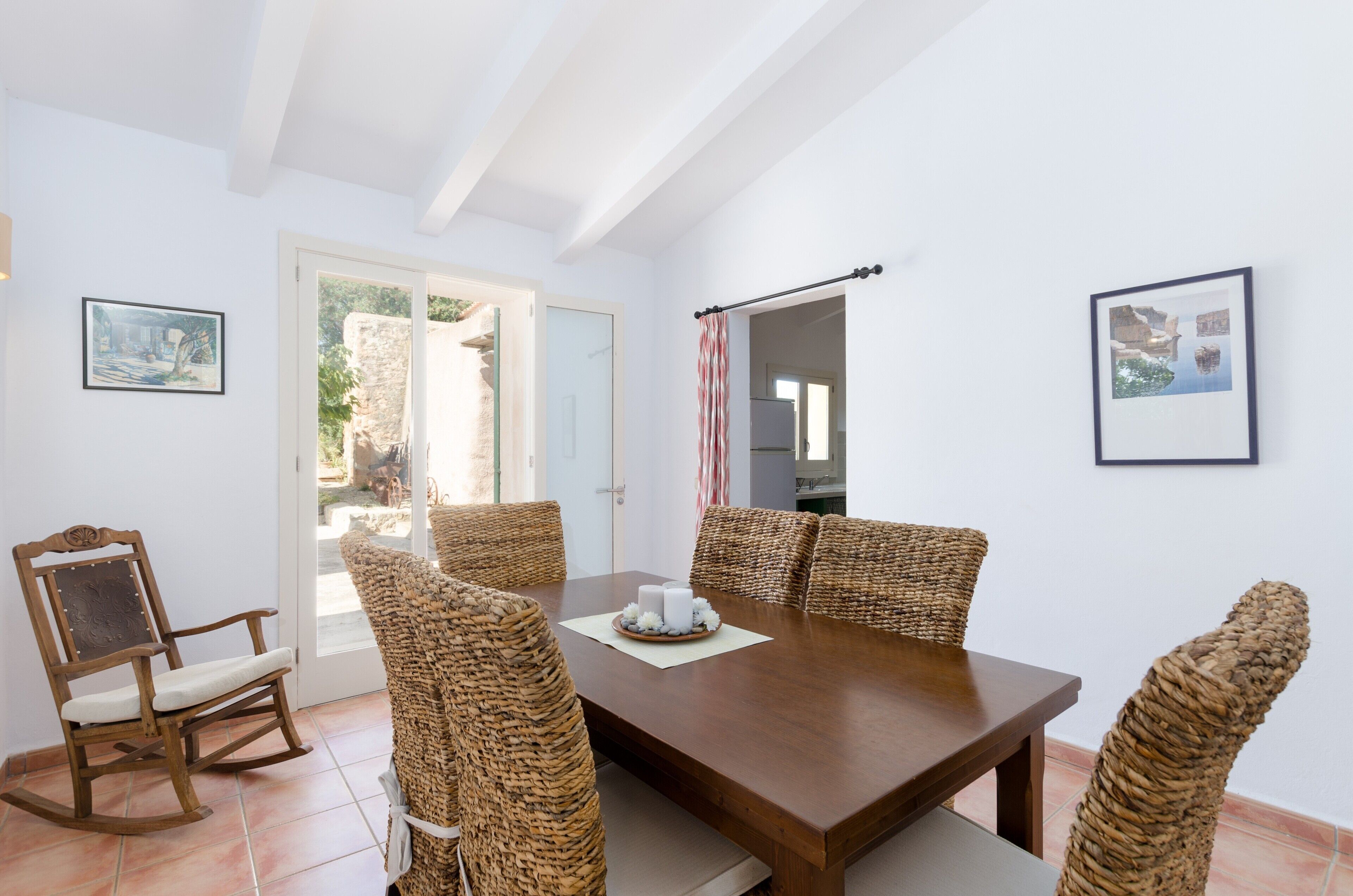 A dining area with a wooden table and wicker chairs, a rocking chair, and framed pictures on the walls.