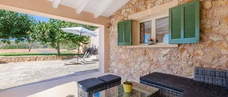 A covered outdoor seating area with wicker furniture, a glass table, and a stone wall with a window and shutters.