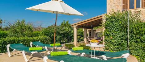 A patio with lounge chairs and a table, surrounded by greenery and a stone building.