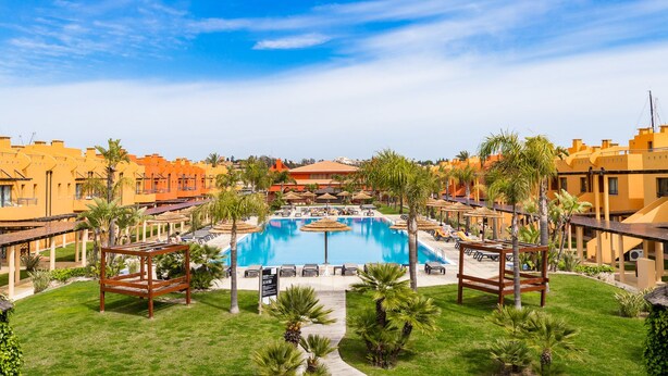 Un resort con piscina central, rodeado de palmeras y sillas de descanso, y edificios al fondo.