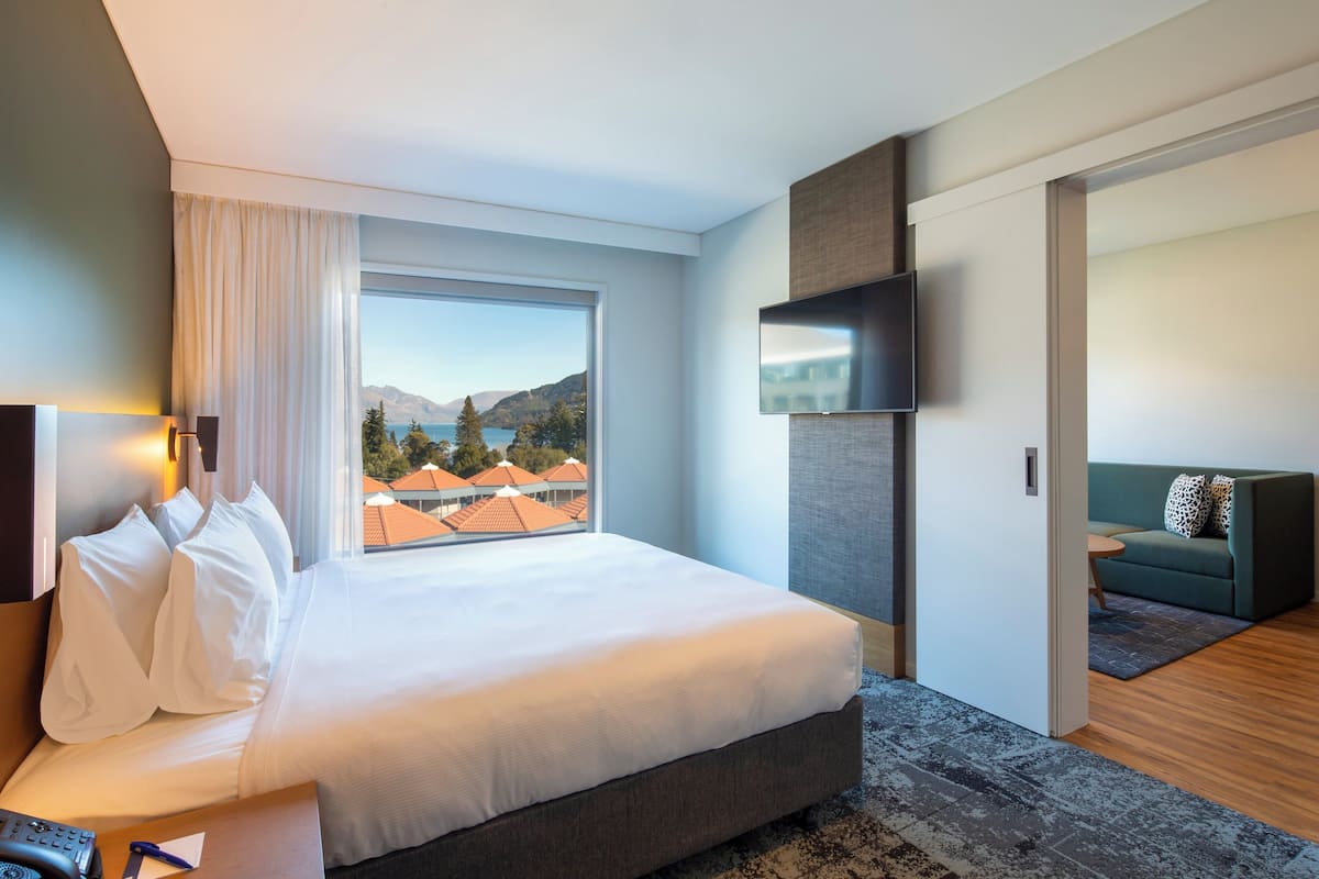 A hotel room with a large bed, a TV mounted on the wall, a view of the mountains and a lake, and a separate seating area with a sofa.