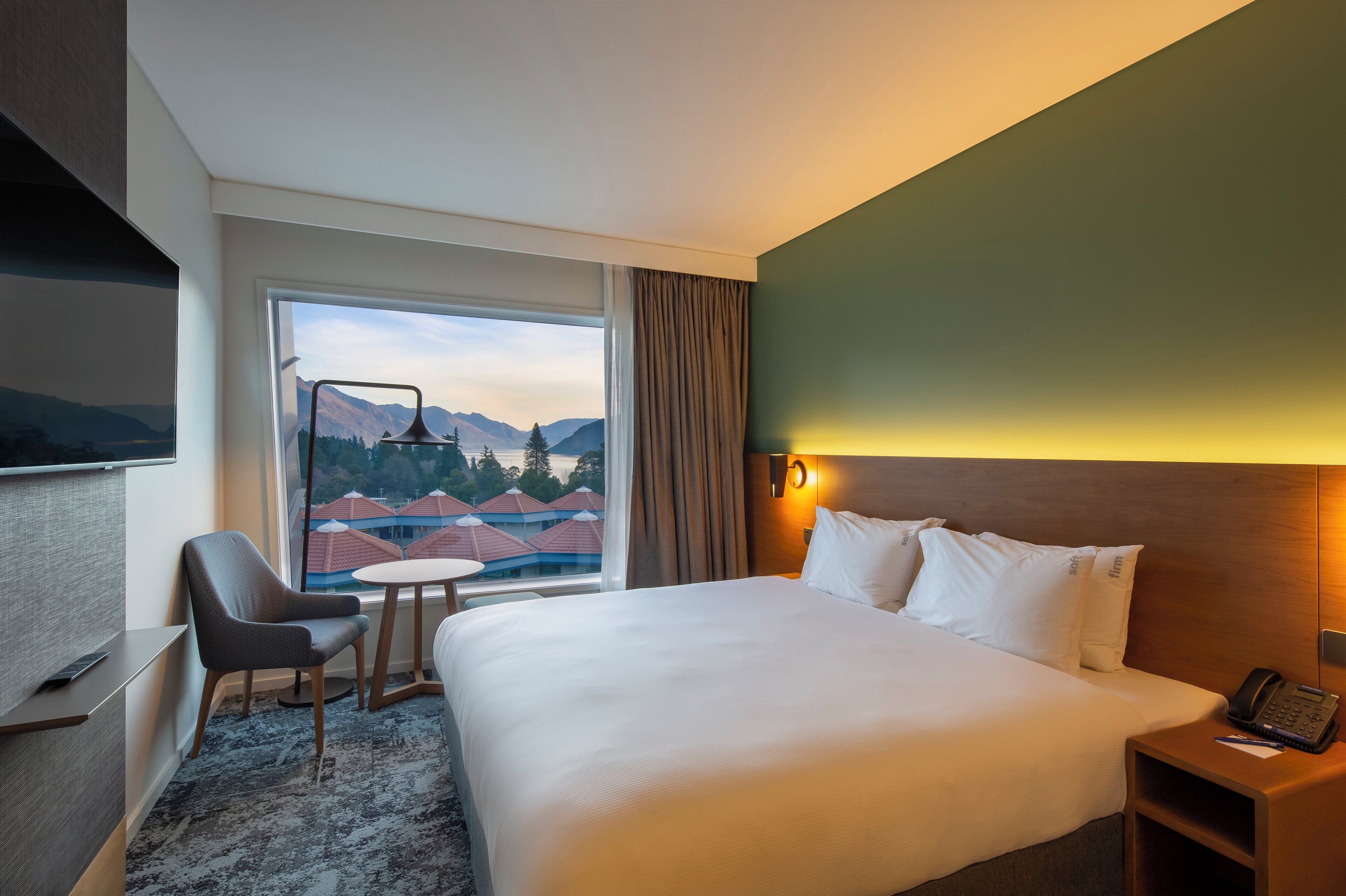 Ett hotellrum med en stor säng, ett skrivbord, en stol, ett litet bord och ett fönster med utsikt över berg och byggnader.