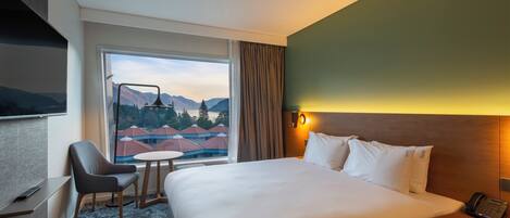 Habitación de hotel con una cama grande, un escritorio, una silla, una mesita y una ventana con vistas a montañas y edificios.