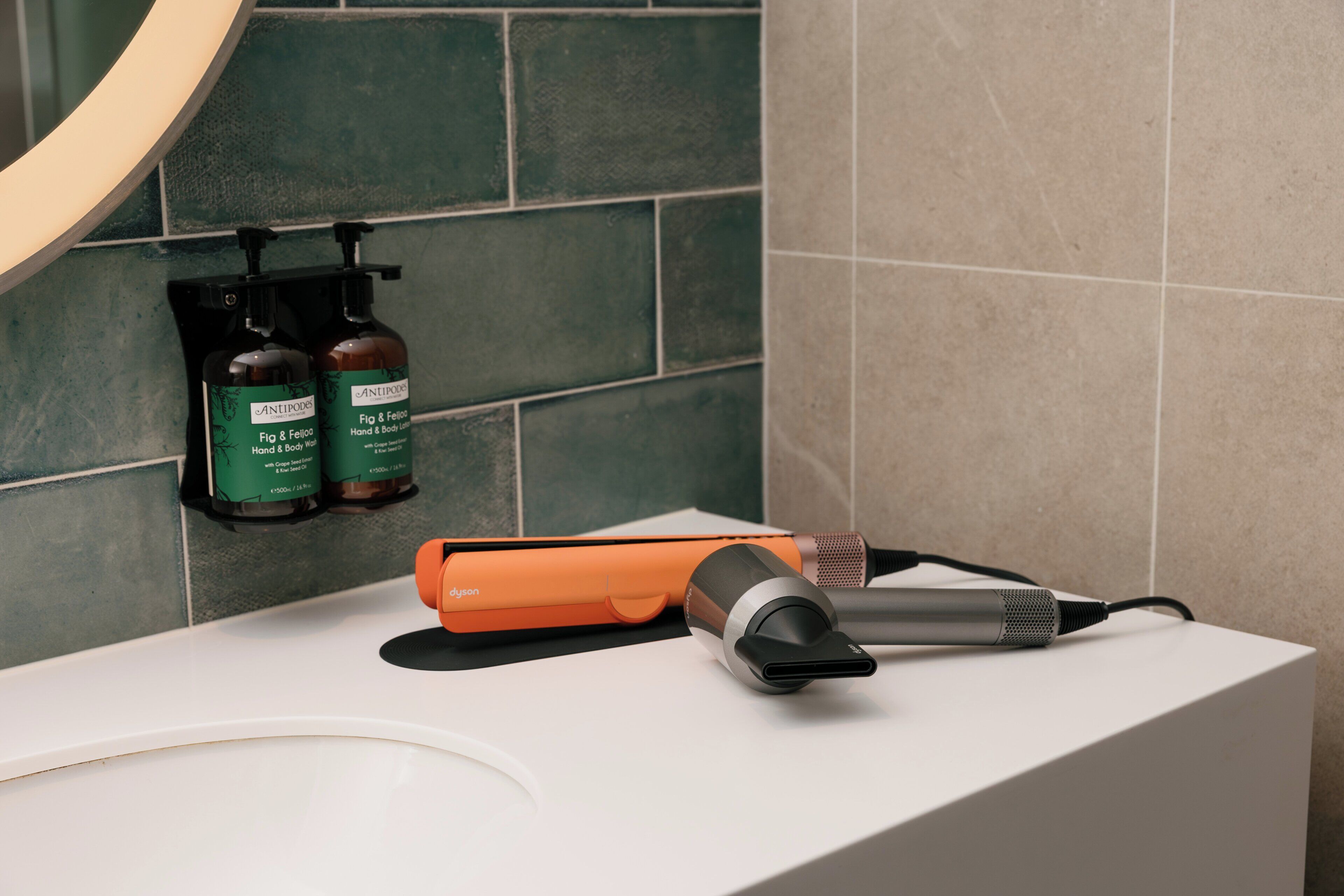 A bathroom with a white sink, a hairdryer, and two bottles of hand and body lotion on a tiled wall.