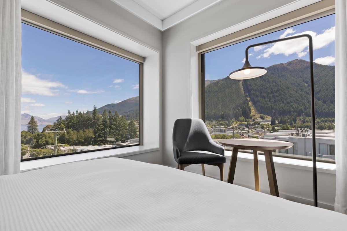 A modern hotel room with a large bed, a chair, a small table, and a lamp. The room has large windows offering a view of mountains and a cityscape.