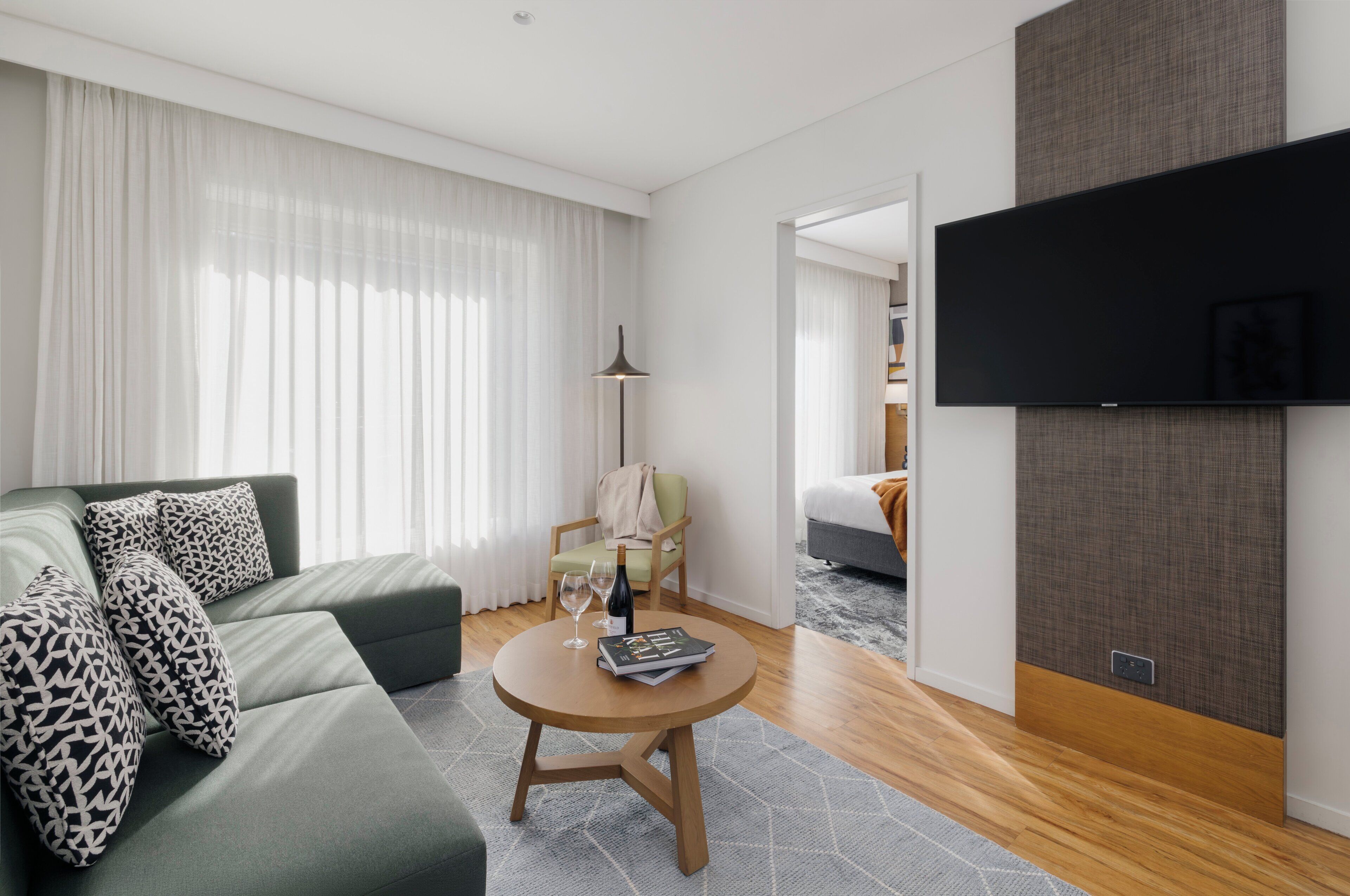 Una habitación de hotel moderna con un sofá verde, una mesa de centro redonda de madera y un televisor de pantalla plana montado en la pared.