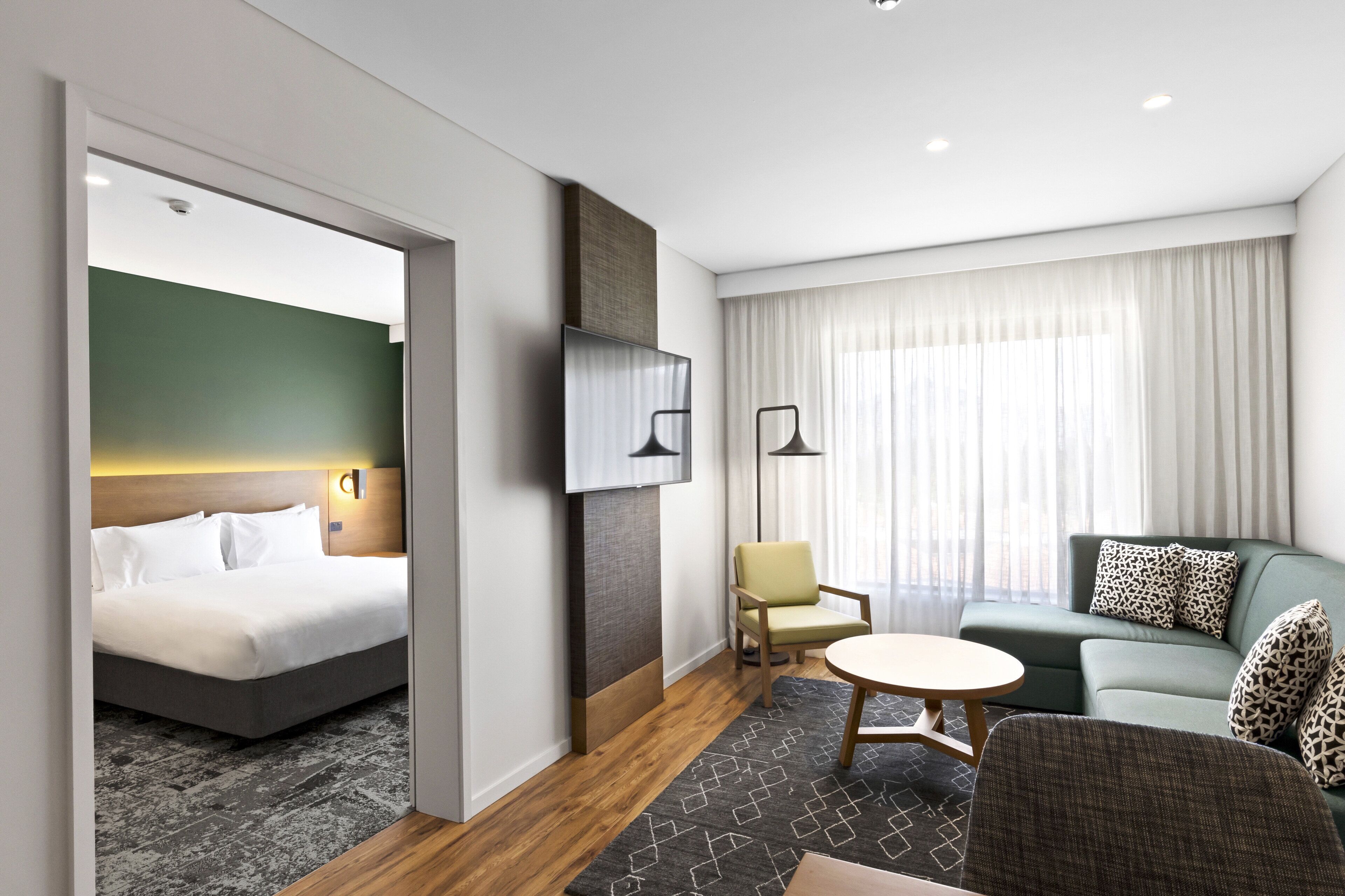 A modern hotel room with a bed, a sofa, a chair, a TV, and a window with curtains.