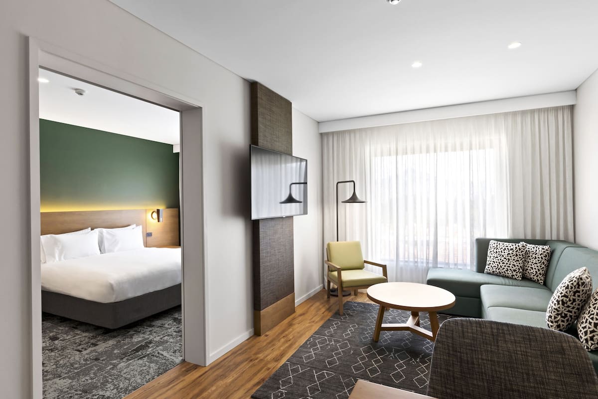 A modern hotel room with a bed, a sofa, a chair, a TV, and a window with curtains.