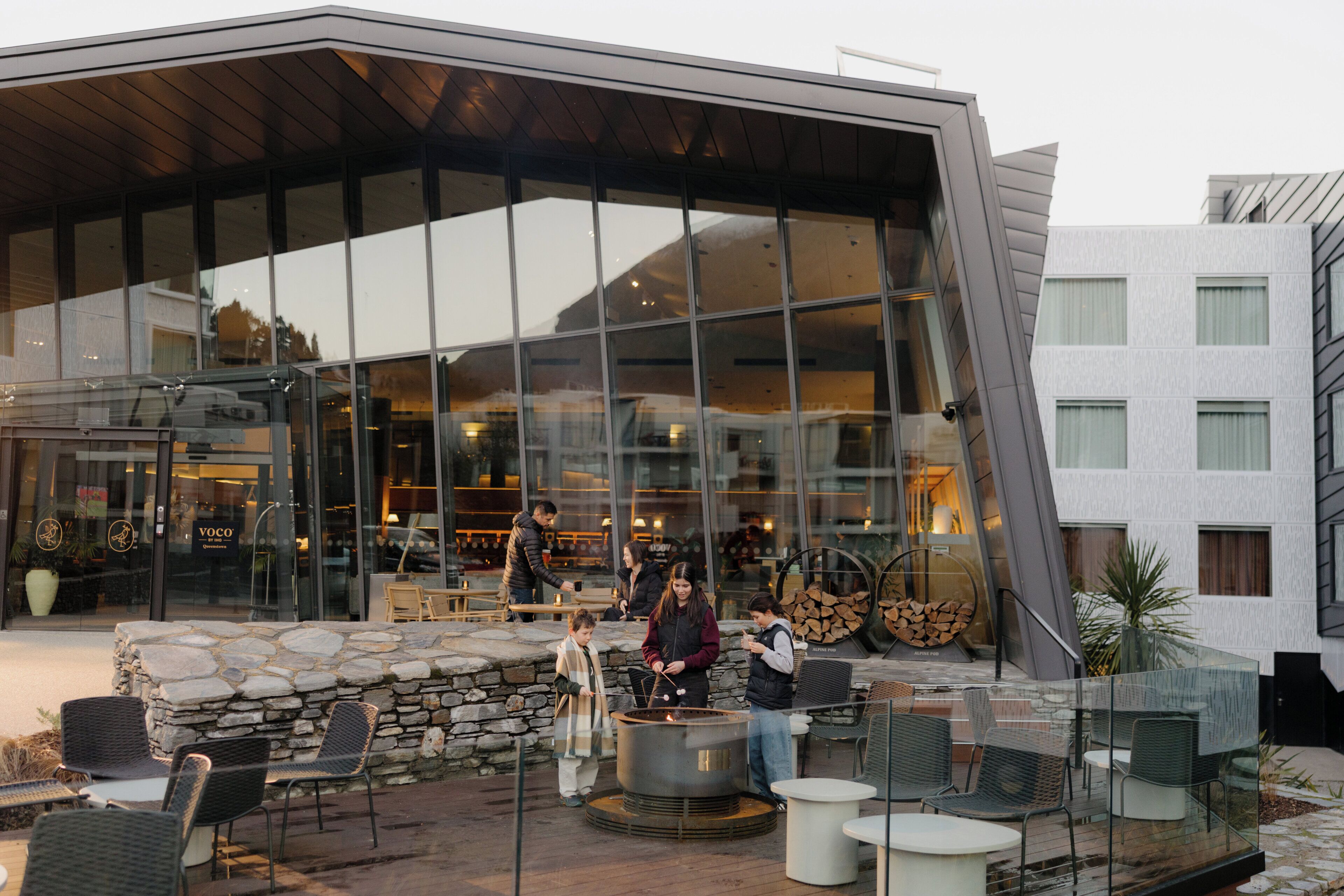 A modern building with a glass facade, outdoor seating area with chairs and tables, and a stone wall.