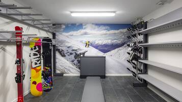 A ski storage room with snowboards and skis on racks, a bench, and a wall mural of a snowy mountain scene.