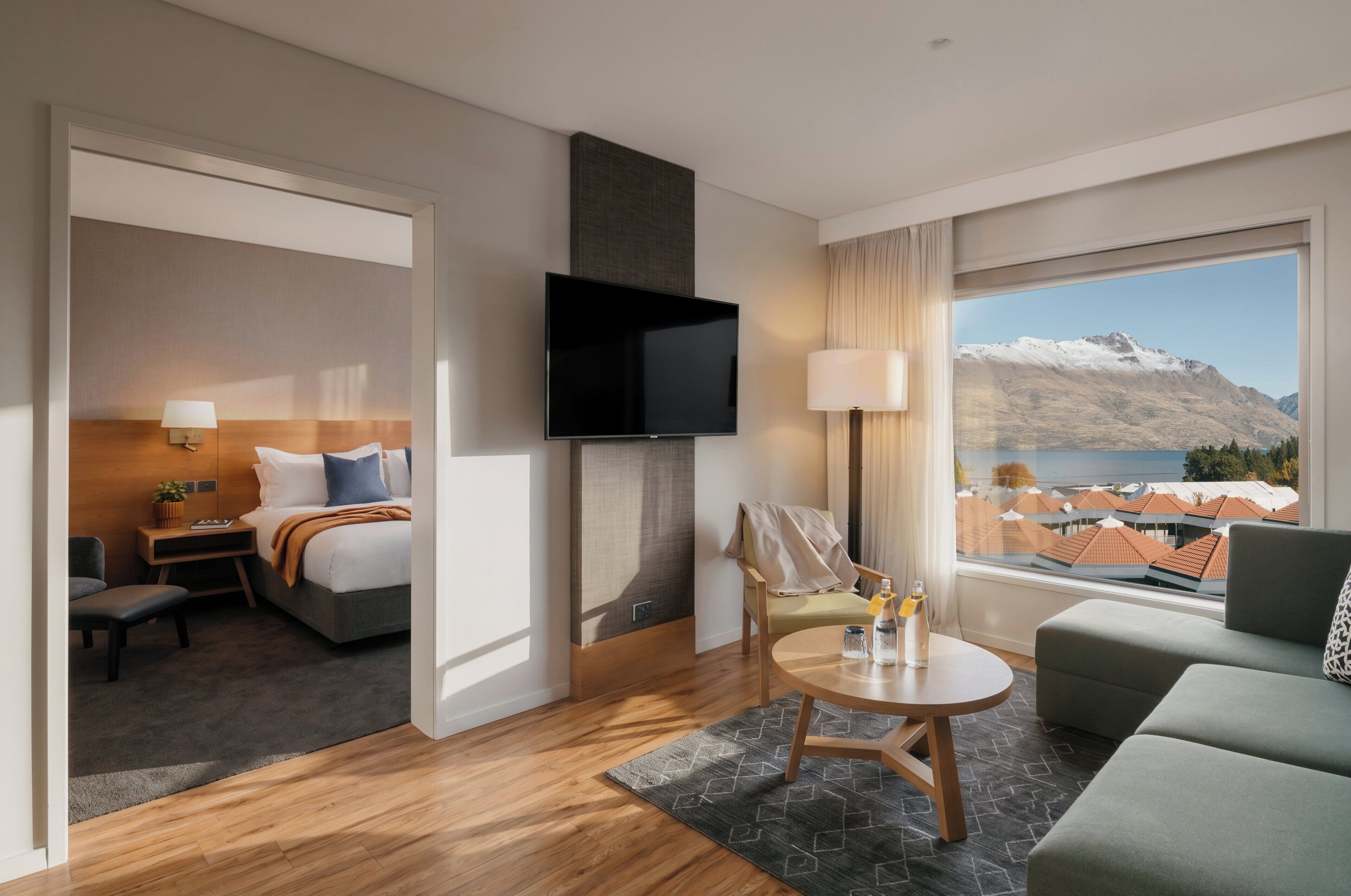 A hotel room with a bed, a TV, a seating area with a table, and a view of mountains through the window.