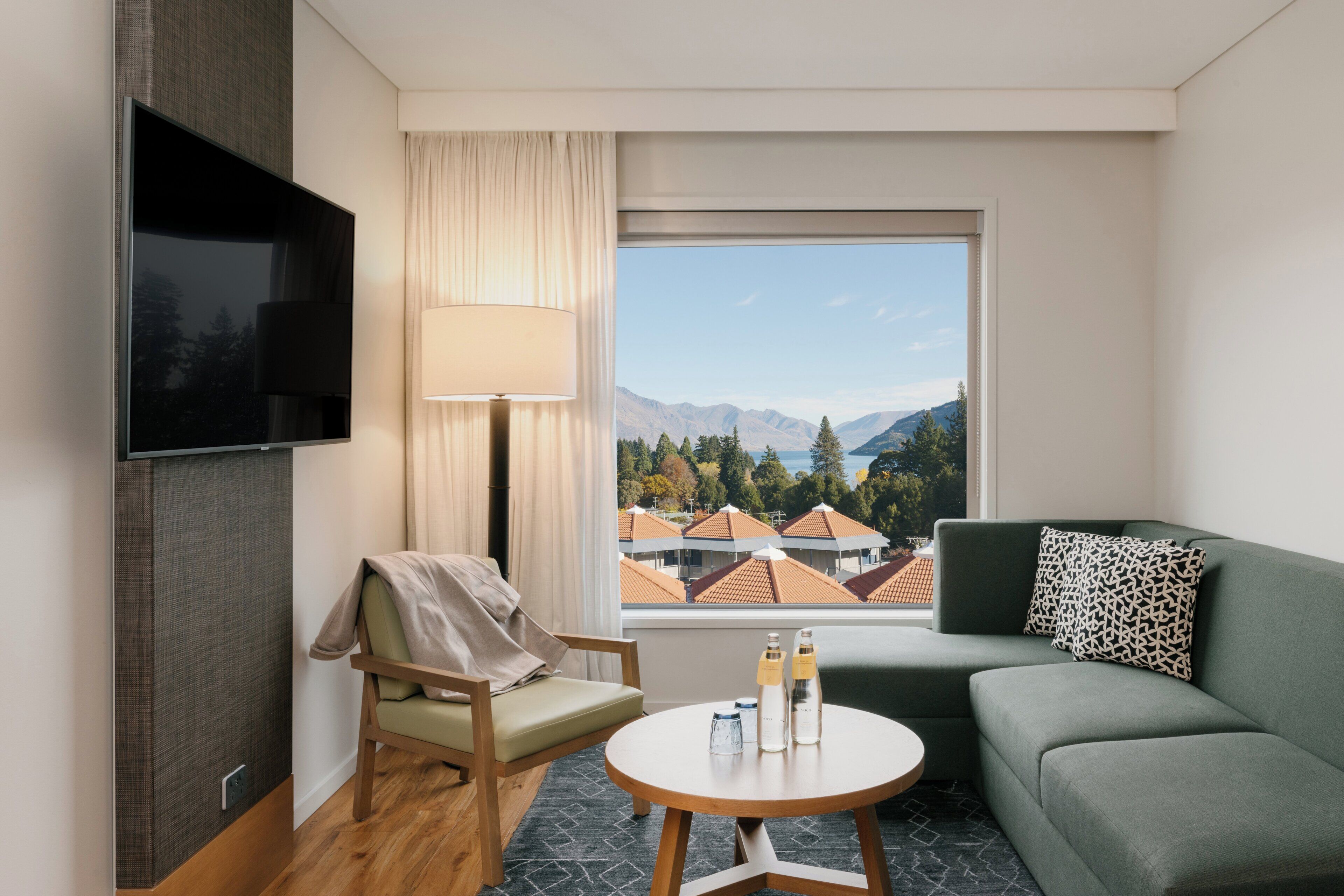 A hotel room with a TV, a sofa, a chair, a table with bottles, and a view of mountains and houses.