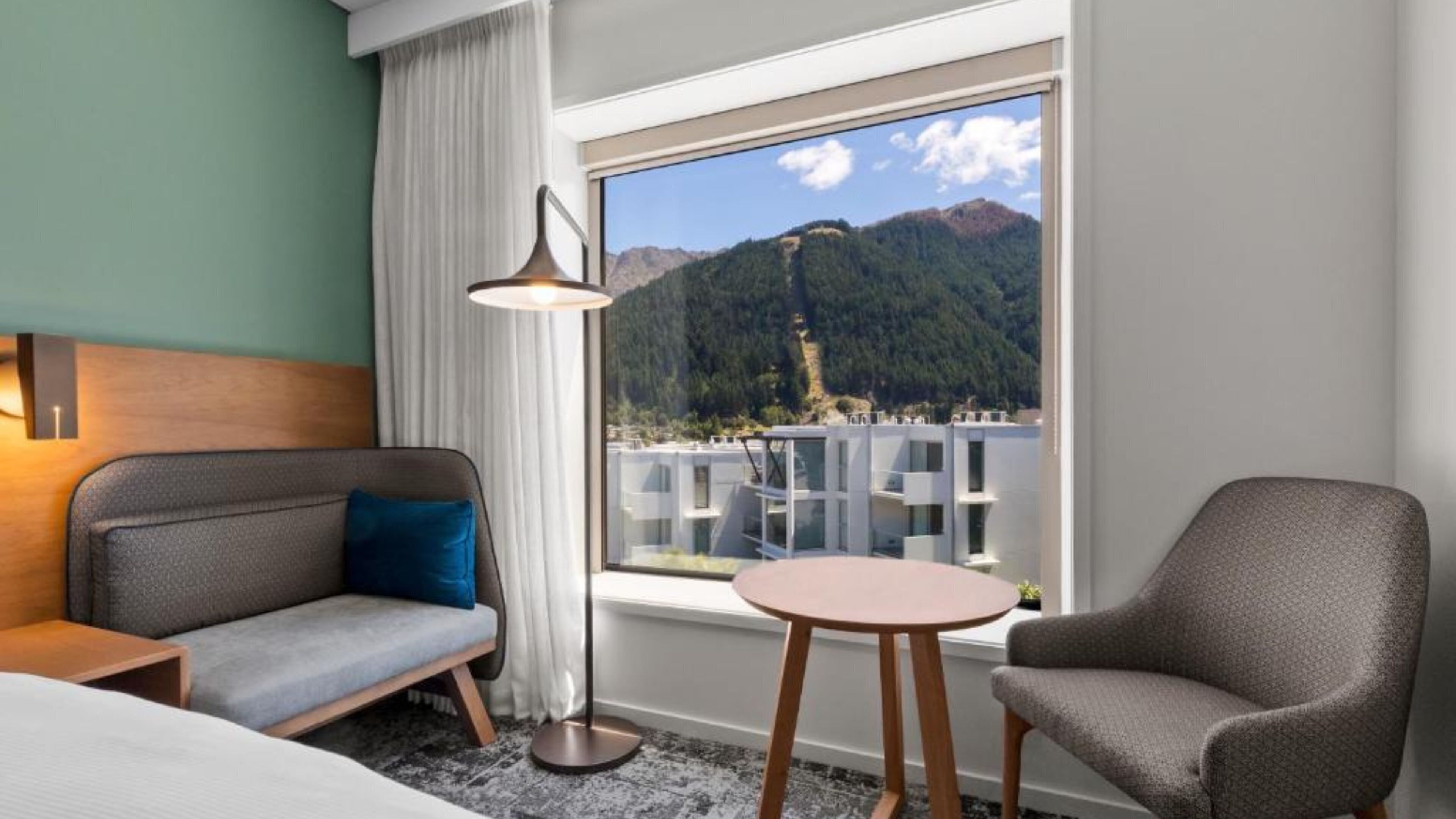 A hotel room with a bed, a sofa, a chair, a small table, a lamp, and a window with a mountain view.