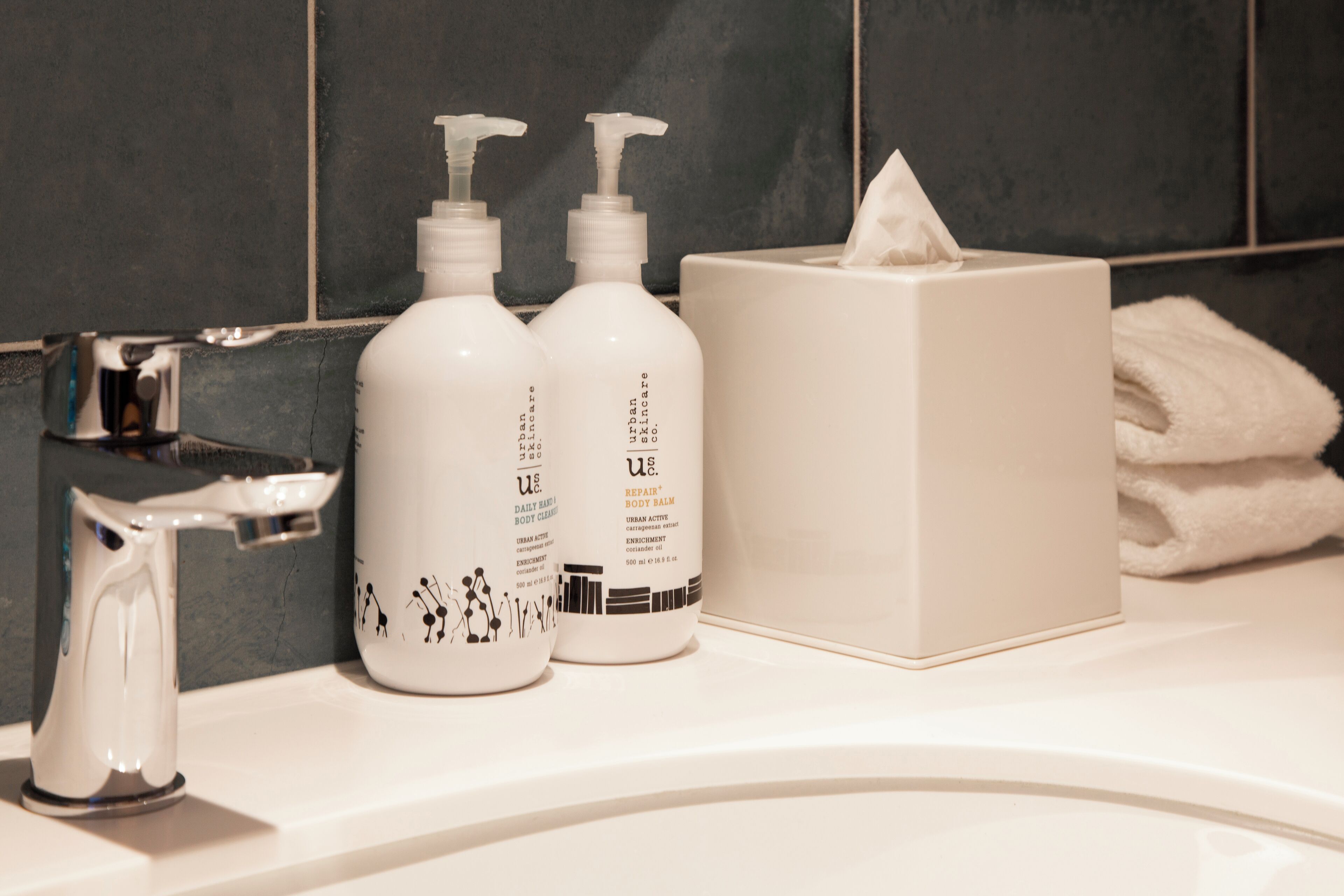 A bathroom sink with two bottles of U.S. Daily Body Care Repair Body Balm, a tissue box, and a towel.