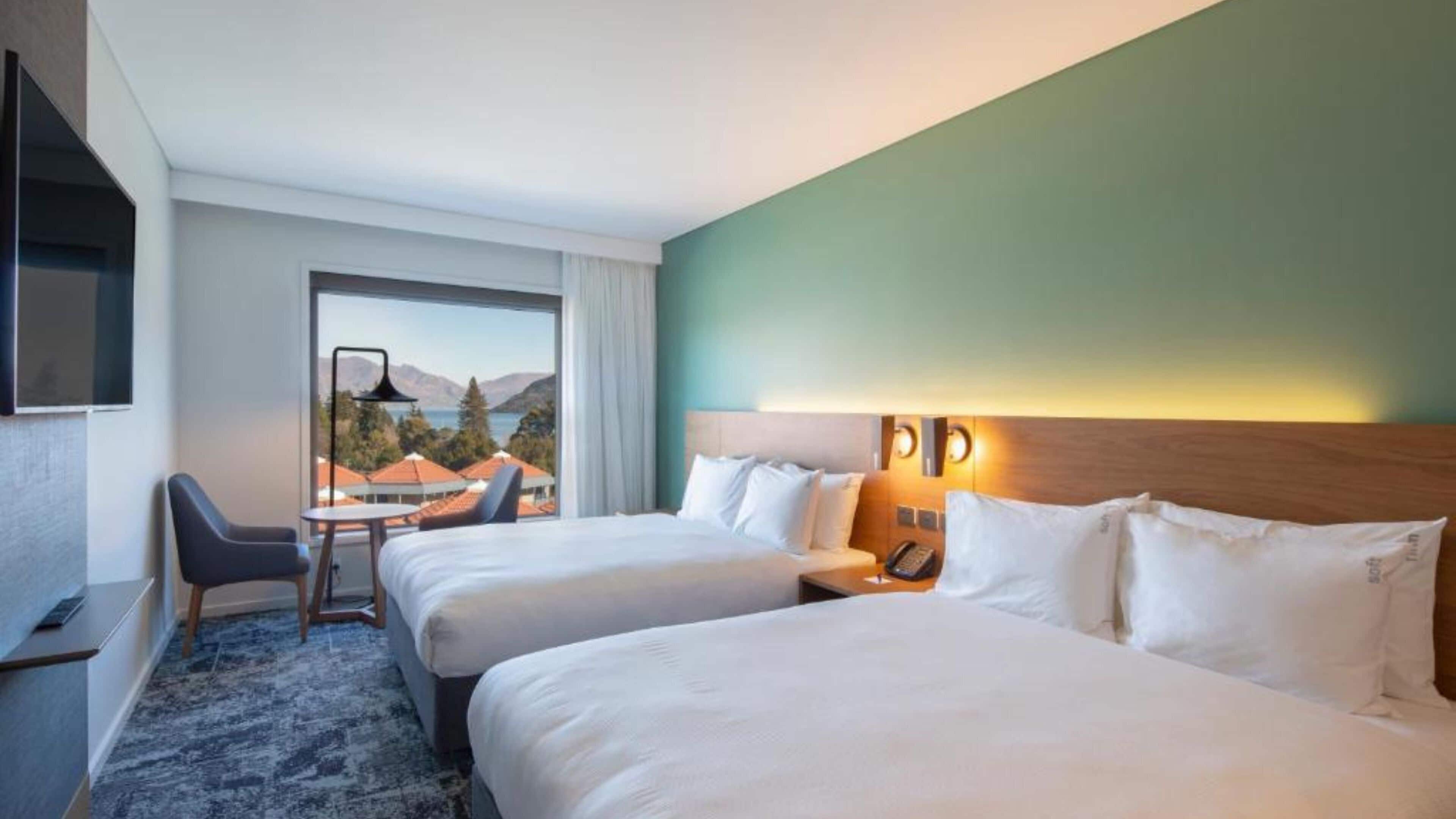 Een hotelkamer met twee bedden, een bureau en een stoel. Er is uitzicht op bergen en een wateroppervlak vanuit het raam.