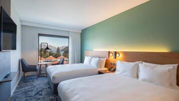 A hotel room with two beds, a desk, and a chair. There is a view of mountains and a body of water through the window.